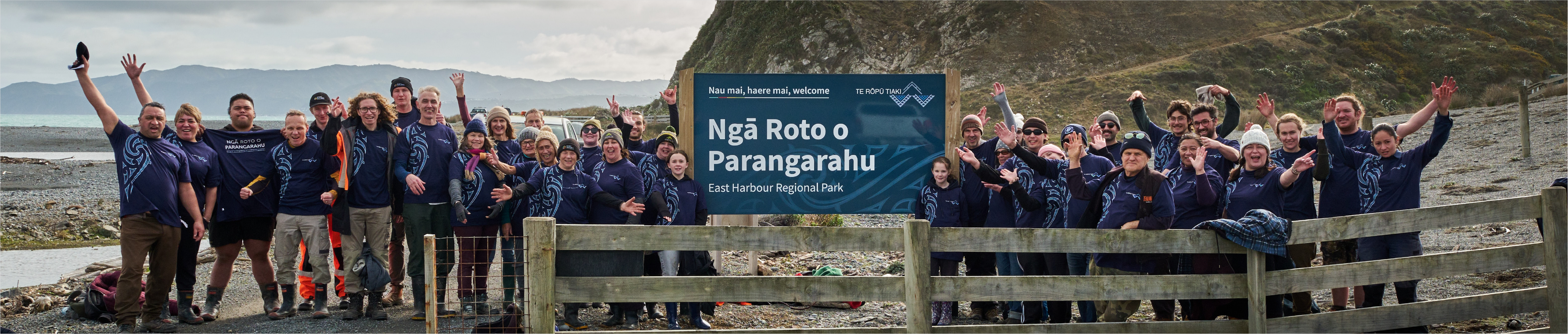 A group of volunteers standing and waving to the camera next to the sign for Parangarahu Lakes at East Harbour Regional Park